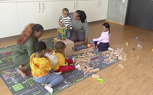 Kinder und Eltern sitzen auf einem Spielteppich und bauen mit Klötzen